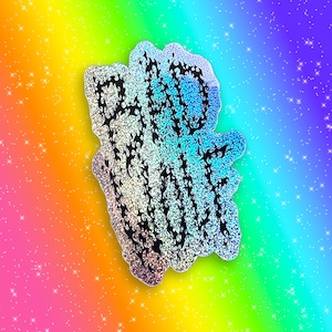 Puede incluir: Una pegatina holográfica con el texto "BAD VIBES" en un diseño de llama negra. La pegatina tiene una forma única y está colocada sobre un fondo degradado de arcoíris con estrellas brillantes.