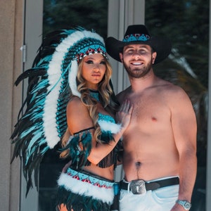May include: A couple dressed in Native American inspired costumes. The woman is wearing a turquoise and black feathered headdress, a turquoise and black beaded top, and a black and white fringed skirt. The man is wearing a black cowboy hat with a turquoise band, white jeans, and a silver belt buckle.
