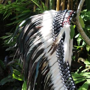 Op de afbeelding: Een witte en zwarte verenhoed met een zwarte en witte gevlochten rand. De hoed heeft een rood en wit parelband met een stervormig ontwerp.