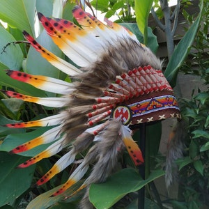 May include: A Native American headdress with red, yellow, and white feathers, a brown leather band with beaded accents, and a red and white beaded design.