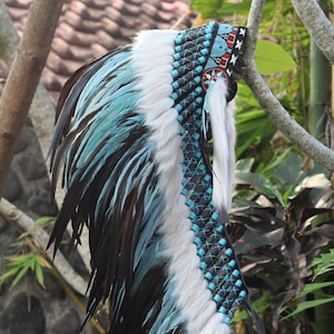 K&ouml;nnte beinhalten: Ein blauer und wei&szlig;er Federkopfschmuck mit schwarzen Lederriemen und Perlen. Der Kopfschmuck ist mit einem rot-wei&szlig;-blauen Muster verziert.