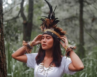 May include: A woman wearing a white dress and a feathered headdress with a beaded necklace. She is standing in a forest with green foliage.