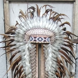 Op de afbeelding: Een traditionele indiaanse hoofdtooi met bruine en witte veren, een kralenband en een leren basis. De hoofdtooi wordt getoond op een houten standaard.