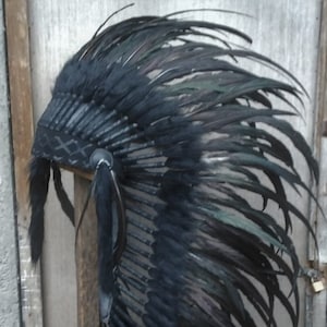 Könnte beinhalten: Ein schwarzer Federkopfschmuck mit einem schwarzen Lederband und einem Perlenmuster. Der Kopfschmuck wird auf einem Holzständer präsentiert.