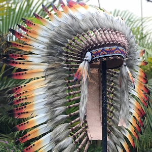 Może przedstawiać: Tradycyjny indiański strój głowy z brązową skórzaną podstawą i paskiem kolorowych koralików. Strój głowy jest ozdobiony białymi i pomarańczowymi piórami z czarnymi kropkami.