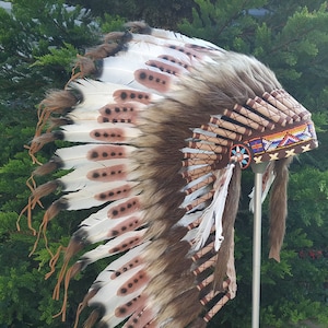 Può includere: Un copricapo nativo americano marrone e bianco con piume, perline e dettagli in pelle. Il copricapo è esposto su un supporto in metallo.