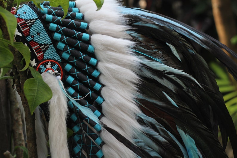 K&ouml;nnte beinhalten: Ein indianischer Kopfschmuck mit wei&szlig;en und blauen Federn, schwarzem und blauem Perlenbesatz und einem roten und blauen Perlenband.