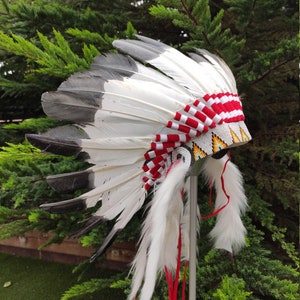 Puede incluir: Tocado nativo americano con plumas blancas y grises, acentuado con bandas tejidas rojas y blancas. La diadema tiene una banda de cuentas con detalles amarillos y negros, y detalles blancos y esponjosos con cintas rojas.