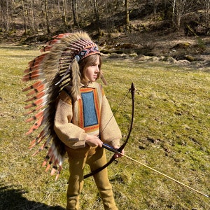 Könnte beinhalten: Ein Kind in einem braunen Pullover und einer beigefarbenen Hose hält einen Bogen und Pfeil. Es trägt ein farbenfrohes indianisches Kopfschmuck mit Federn. Das Kind steht auf einer grasbewachsenen Wiese mit Bäumen im Hintergrund.