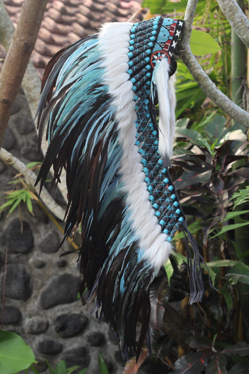 K&ouml;nnte beinhalten: Ein blauer und wei&szlig;er Federkopfschmuck mit schwarzen Lederriemen und Perlen. Der Kopfschmuck ist mit einem rot-wei&szlig;-blauen Muster verziert.