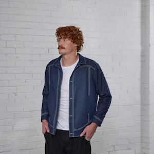 Op de afbeelding: Een blauw denim jack met witte stiksels en een wit T-shirt eronder. De persoon draagt een zwarte broek.