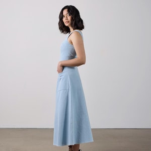 May include: A light blue sleeveless dress with a flared skirt and large front pockets. The dress is paired with black strappy sandals. The model is standing in front of a white wall.