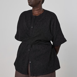 May include: Black button-down top with a textured pattern and a matching belt. The top has short sleeves and is paired with brown pants. The outfit is a modern take on a classic style.