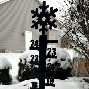 May include: A black metal snow depth gauge with a snowflake at the top and numbers from 4 to 24 inches. The gauge is standing in a snowdrift.