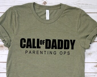 May include: Olive green t-shirt with the text "CALL OF DADDY PARENTING OPS" in bold black letters. The shirt has a crew neck and short sleeves, and is made of a soft, comfortable material.