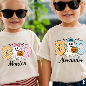 May include: Two off-white t-shirts featuring a graphic of a ghost holding a pumpkin with the text BOO. One shirt displays the name Monica with a pink bow on the ghost, and the other displays the name Alexander with a blue baseball cap on the ghost.