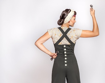 May include: A woman wearing a floral print top and dark green overalls is holding a wrench in each hand. She is standing in front of a spare tire and a wooden toolbox.