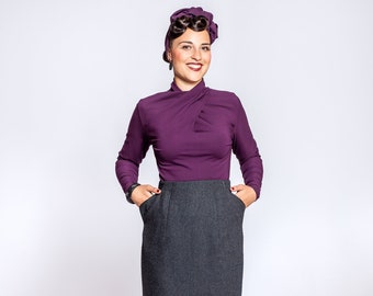 May include: A woman wearing a purple long-sleeved blouse and a dark gray pencil skirt. She is standing with her hands in her pockets and is smiling at the camera.