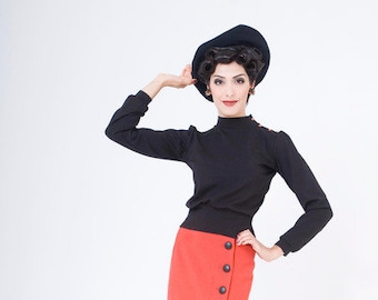 May include: A woman wearing a black turtleneck sweater and a red skirt with buttons down the side. She is wearing a black hat and black heels. She is standing next to a vintage suitcase.