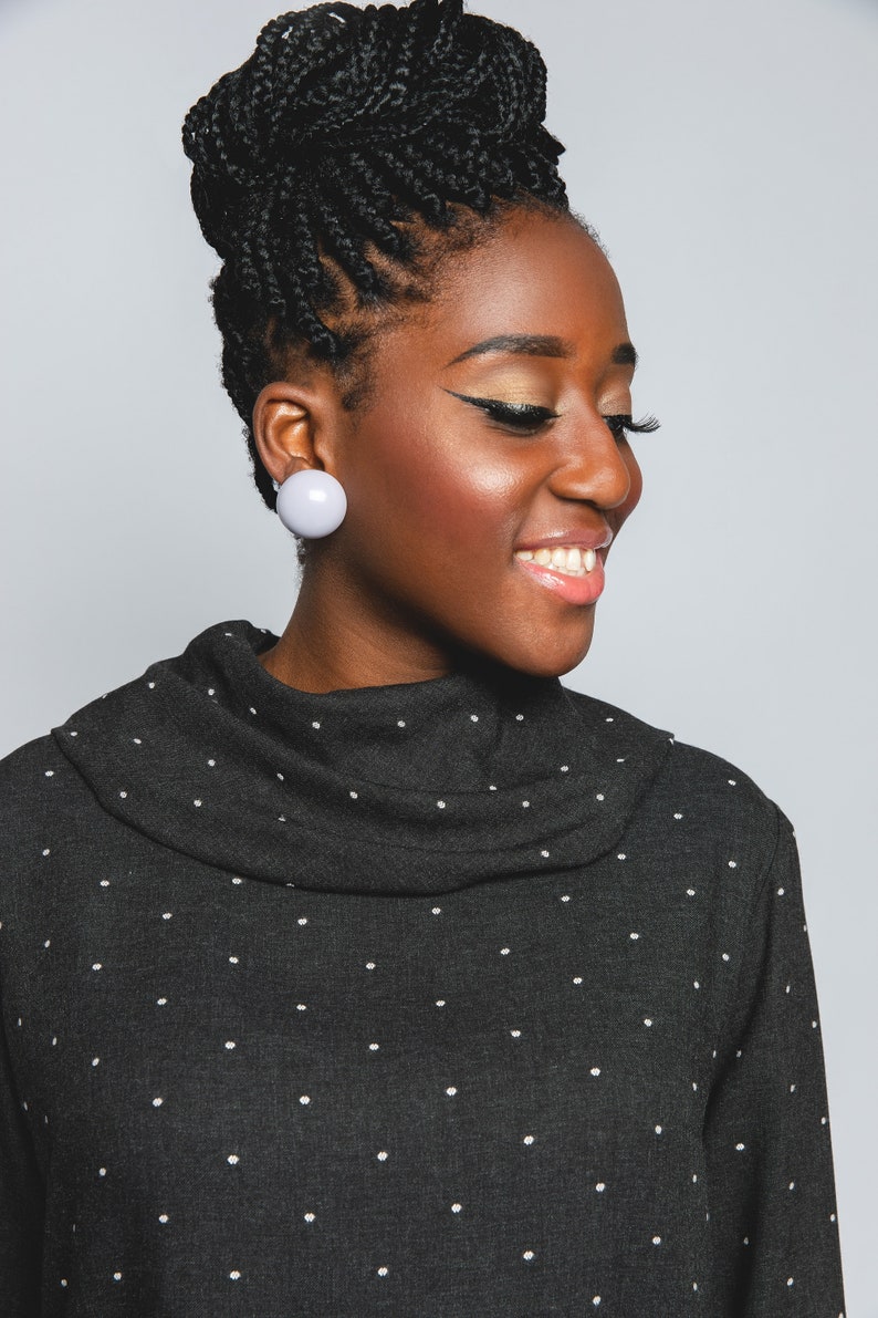 May include: A woman with black braids and a black turtleneck sweater with white polka dots. She is wearing large, round, light purple earrings.