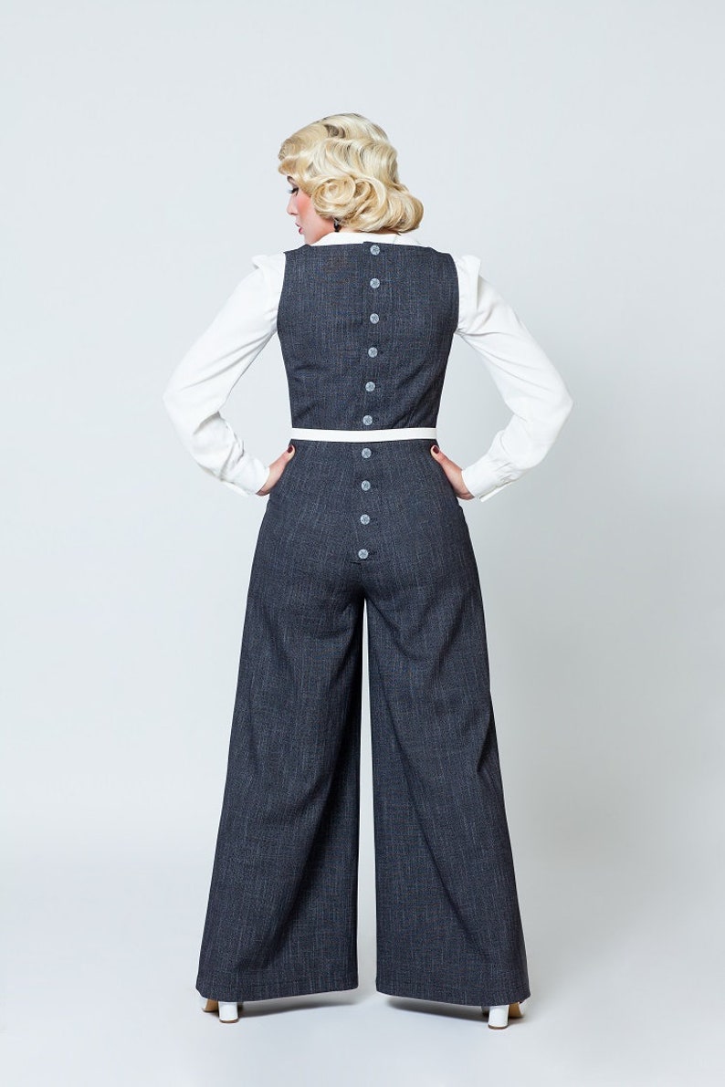 May include: A woman wearing a dark gray wide-leg jumpsuit with a white trim and button detailing on the back. She is wearing a white blouse underneath the jumpsuit.