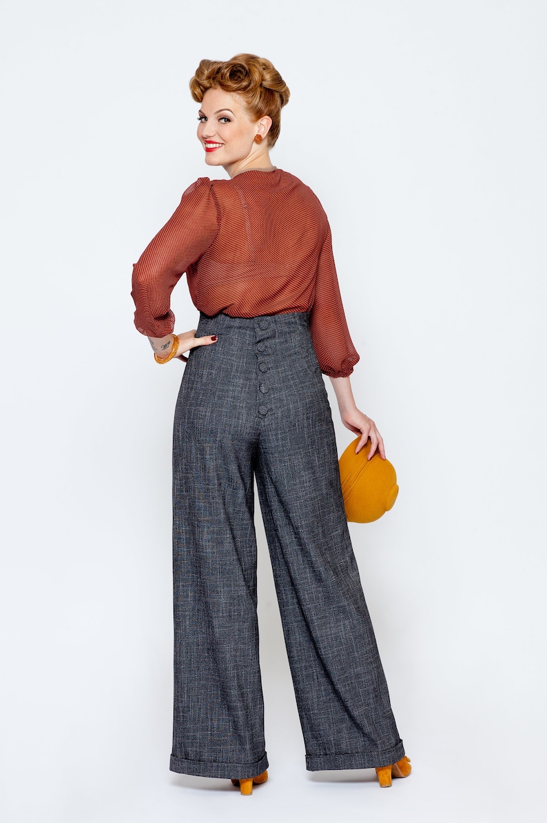 May include: A woman wearing a rust-colored blouse and high-waisted wide-leg pants with a button detail down the back. She is holding a mustard yellow hat.