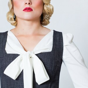 May include: A woman wearing a white blouse with a white bow tie and a gray pinstripe vest.