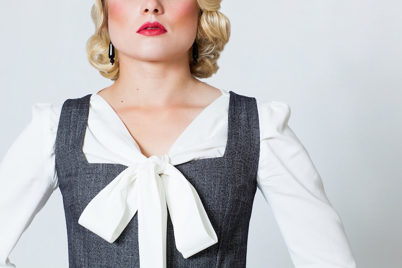 May include: A woman wearing a white blouse with a white bow tie and a gray pinstripe vest.