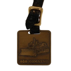 May include: A vintage Komatsu key tag. The bronze-coloured rectangular plate features an embossed bulldozer design. It's attached to a black leather strap with a gold-coloured buckle. The Komatsu logo is at the bottom of the tag.