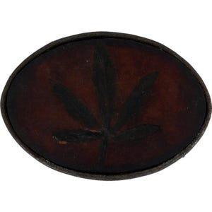 May include: Oval-shaped belt buckle with a dark brown leather background. A black cannabis leaf is painted on the leather. The buckle has a dark, textured metal border.