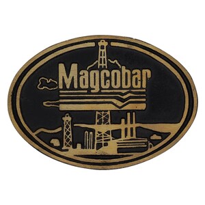 Puede incluir: Un emblema ovalado de metal negro y dorado con la palabra "Magcobar" en letras doradas. El emblema presenta una ilustración de una plataforma petrolífera y otras estructuras industriales.