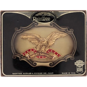 May include: A rectangular Harley-Davidson belt buckle with a gold eagle design. The eagle is centered on a gold background, with a red banner below that reads "HARLEY-DAVIDSON". The buckle is black with the text "RainTree" at the top and "Made in U.S.A." at the bottom.
