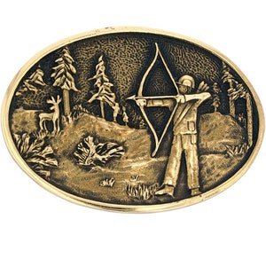 May include: Oval-shaped, gold-toned belt buckle featuring a detailed relief scene. The design depicts an archer aiming a bow and arrow in a forest setting, with trees, a deer, and foliage. The buckle has a textured background and a polished border.