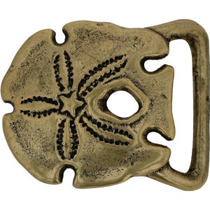 May include: A brass-colored belt buckle in the shape of a sand dollar. The buckle has a detailed design with a central star and radiating lines. The buckle has a rectangular loop for the belt.