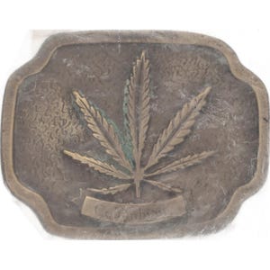May include: A rectangular, bronze-colored belt buckle featuring a cannabis leaf design. The leaf is detailed with gold and green accents, and the word "Columbia" is inscribed on a banner below the leaf.