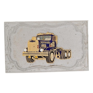 Puede incluir: Un objeto rectangular plateado con un borde ornamentado. El centro presenta una ilustración detallada de un camión Autocar azul y dorado. El camión tiene neumáticos negros y la palabra "Autocar" es visible.