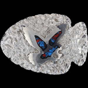 Könnte beinhalten: Gürtelschnalle aus silberfarbenem Metall in Pfeilspitzenform. Sie zeigt ein stilisiertes Vogeldesign mit roten und blauen Steineinlagen. Der Hintergrund hat ein strukturiertes, gehämmertes Aussehen.