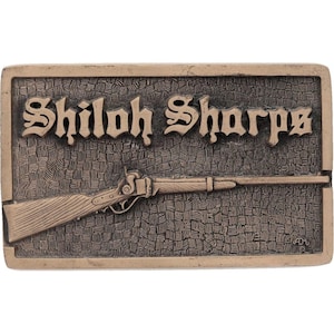 May include: A rectangular, bronze-colored belt buckle featuring the words "Shiloh Sharps" in an ornate font above a rifle illustration. The background has a textured pattern. The buckle is likely made of metal.