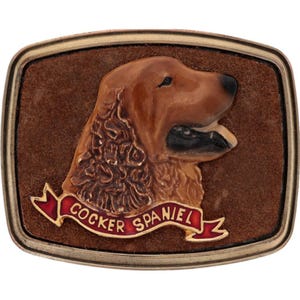 May include: A rectangular belt buckle featuring a brown Cocker Spaniel dog in profile. The dog is set against a brown background with a gold-colored border. A red banner with the words "COCKER SPANIEL" is at the bottom.
