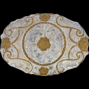 May include: An oval silver belt buckle with detailed floral and scroll patterns. Gold-coloured accents highlight the design, including flowers and a rope-style border. The background is a dark black.