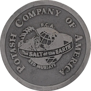 Op de afbeelding: Een ronde, grijze metalen medaillon met verhoogde letters en afbeeldingen. De tekst luidt "POTASH COMPANY OF AMERICA" rondom. Het midden toont een Indiaans profiel, een wereldbol en de woorden "The SALT of the EARTH" en "RED MURIATE".