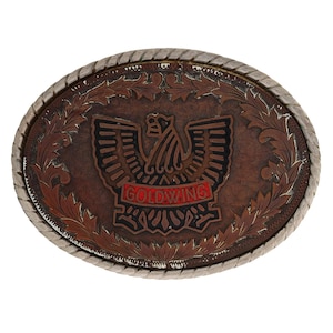 May include: Oval-shaped belt buckle with a brown leather-like center featuring an eagle design and the word "GOLDWING" in red. The buckle has a silver-colored rope-like border and decorative leaf patterns.
