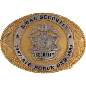 Può includere: Una fibbia per cintura in oro e argento con il testo "BMAC SECURITY 1987-AIR FORCE ONE-1989" e un design stilizzato di aquila e scudo.
