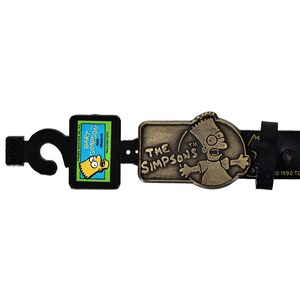 May include: A black leather belt with a metal buckle featuring Bart Simpson from The Simpsons. The buckle has the text "THE SIMPSONS" and "TM". The belt also has a Bart Simpson graphic and the text "BART SIMPSON AND GROEONG".