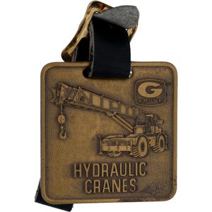 Peut inclure: Une étiquette métallique de couleur bronze avec une lanière en cuir noir. L'étiquette présente une image en relief d'une grue hydraulique et les mots "HYDRAULIC CRANES" et le logo "GROVE".