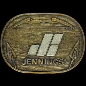 May include: A brass belt buckle with a silver Jennings logo and a bow and arrow design. The buckle has a textured surface and a registered trademark symbol.