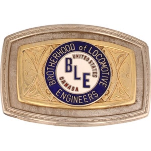 May include: A gold and silver belt buckle with a blue and white circular emblem that reads "Brotherhood of Locomotive Engineers" and "United States Canada" with the letters "BLE" in the center.