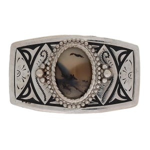 Può includere: Fibbia rettangolare color argento con un centro in pietra ovale e macchiata. La fibbia presenta intricati disegni geometrici neri e argento e un bordo di perline attorno alla pietra. Il design generale è dettagliato e ornato.