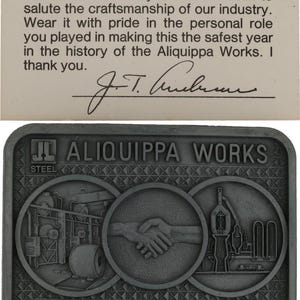 May include: A rectangular, grey metal buckle with the text "ALIQUIPPA WORKS" and "SAFEST YEAR 1980." The buckle features three circular designs with industrial imagery and a handshake. The text above the buckle reads, "The die for this limited edition buckle..."