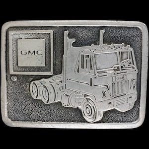 May include: A silver belt buckle featuring a detailed illustration of a GMC semi-truck. The buckle has the GMC logo on the left side and the word "GMC" on the front of the truck.